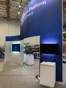 RENTAL: Modified RE-9135 Island Design with Curved Gravitee Panel System Wall, (5) RE-1575 White Laminated Workstation Counters, (1) RE-1576 White Laminated Reception Counter, (6) Monitor Mounts, (5) 43" Monitors, (1) 65" Monitor, (8) LED Arm Lights, 10 f